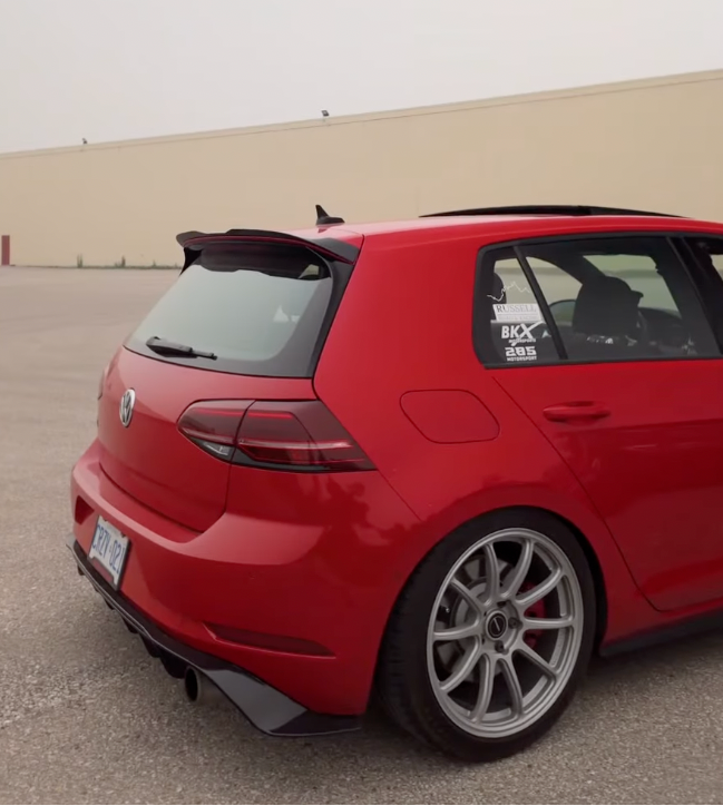 A red Volkswagen Golf R hatchback parked outdoors on a paved lot, viewed from the rear three-quarter angle. The car features a black rear spoiler, custom silver multi-spoke wheels, and a tinted rear window with a visible 'R33' decal.